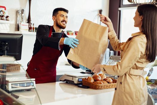 Shop assistant handling shopping bag to female customer in grocery store, close up