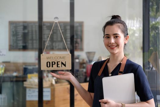 Beautiful asian young barista woman in apron holding tablet and standing in front of the door of cafe with open sign board. Business owner startup SME entrepreneur concept