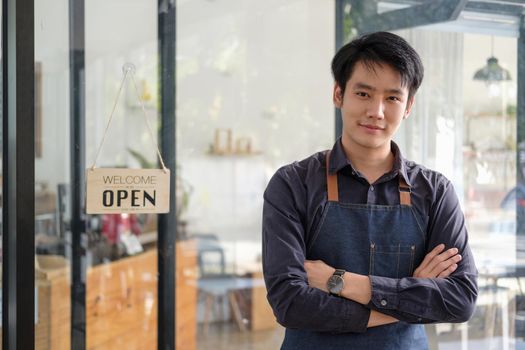 Smart asian young barista male in apron holding tablet and standing in front of the door of cafe with open sign board. Business owner startup SME entrepreneur concept.