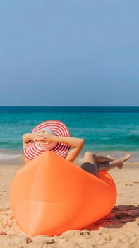 Summer lifestyle portrait of pretty girl sitting on the orange inflatable sofa on the beach of tropical island. Relaxing and enjoying life on air bed. VERTICAL FORMAT for Instagram mobile story or stories size. Mobile wallpaper