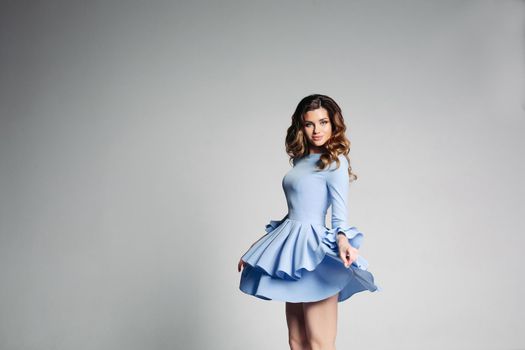 Portrait of beautiful young woman with long curly hair and shining blue eyes, wearing in fashionable light blue dress with flounces over grey studio background. Dress rose from the movement of girl.
