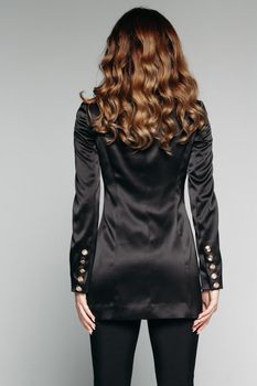 Back view of unrecognizable model with brunette wavy hair of medium length wearing casual black jacket and trousers made from silk. Isolate on grey. Studio shot.