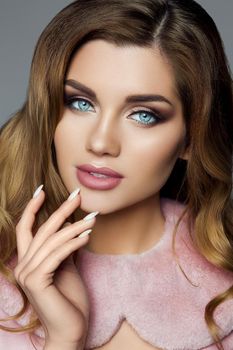 Studio portrait of attractive wavy haired young woman with blue eyes and bright professional make-up holding hand with manicure wearing pink velvet collar over grey background.