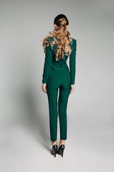 Back view of faceless blonde woman with hairstyle wearing skinny trousers and green jackets with heels over white background.