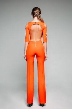 Full length back view of fashionable model in casual bright orange overall with open back and high heels over grey background.