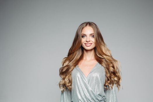 Front view of seductive and stylish blonde woman with volumed curly hair, perfect make up after beauty salon smiling at camera. Pretty positivity girl wearing in gray shiny blouse. Gray background.