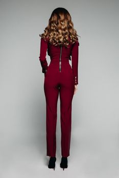 Back view of slim model with long brunette hair wearing bordo suit and black high heels. Cutout on grey background.