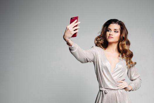 Stylish and attractive woman confident posing with hand on hips and taking self portrait at call phone. Sudactive female model in silver dress taking photo at smartphone. Concept of style.