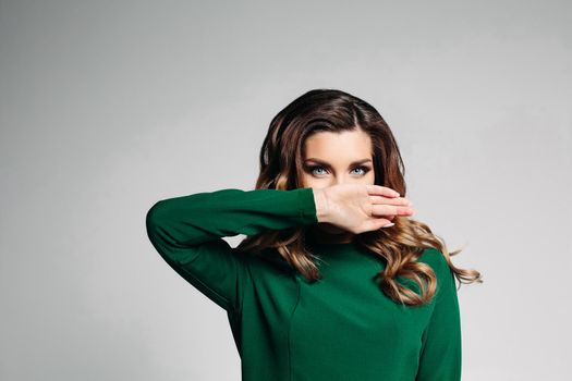 A beautiful brunette in a green dress with blue eyes and curly hair, looks into the camera and gently covering her mouth with her right hand. Isolate on a gray background