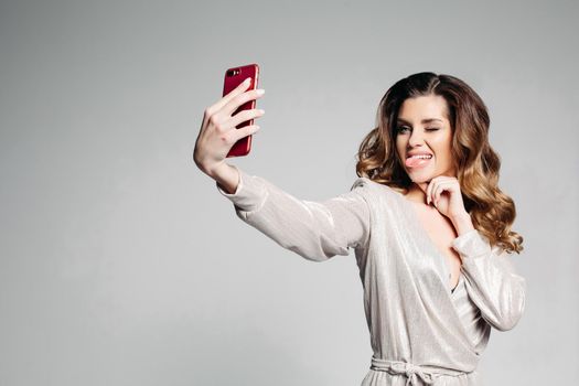 Portrait of a brunette woman dressed in a stylish gray dress, with make-up and hairstyle, makes selfie showing her tongue and fisting her eyes, for social networks. Bright girl isolated