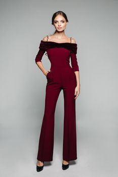 Full length of blonde model with long wavy hair in tail posing in fashionable elegant overall of bordo color with fur rim and bare shoulders, wearing high heels.