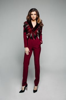 Full length portrait of stylish model with brunette wavy hair wearing elegant bordo overall with long trousers and sleeves with decorated neck area, wearing black high heels. Isolate on grey.