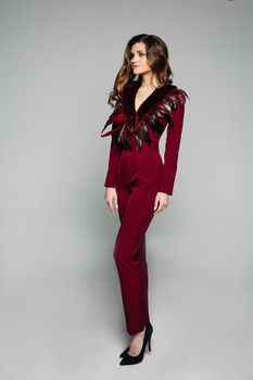 Full length portrait of stylish model with brunette wavy hair wearing elegant bordo overall with long trousers and sleeves with decorated neck area, wearing black high heels. Isolate on grey.