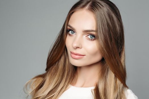 Studio portrait of an attractive blond woman with long thick hair and low-key makeup, wearing an elegant white blouse, cute looks at the camera on a gray background. Isolate. The concept of beauty.