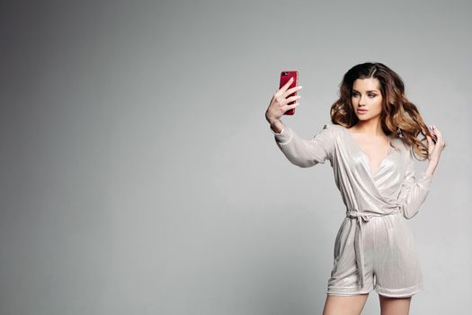 Stylish and attractive woman confident posing with hand on hips and taking self portrait at call phone. Sudactive female model in silver dress taking photo at smartphone. Concept of style.