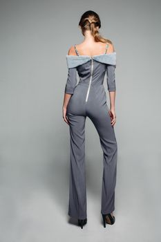 Back view of trendy model wearing elegant grey overall with long trousers and bare shoulders decorated fur and jewelry wearing high heels.