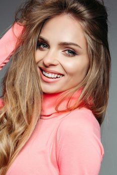 Portrait of pretty and gorgeous blonde woman in pink sweater with wavy volumed hair, sensuality smiling at camera. Girl with perfect make up after beauty salon. Gray studio background.