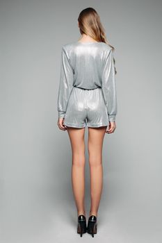Back view full length of unrecognizable model with long legs wearing trendy silver overall and black high heels against grey background. Isolate.