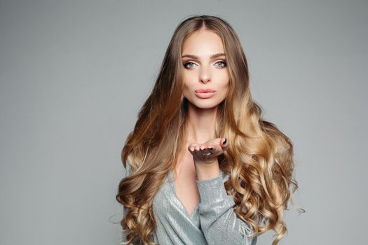 Studio portrait of an attractive blond woman with long thick hair and professional make-up wearing an elegant silver blouse, an air kiss on the camera on a gray background. Isolate. The concept of beauty.