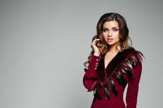 Stunning model with brunette wavy hair and blue eyes looking at camera in bordo overall decorated with fur and feathers. Isolate on grey. Copyspace.
