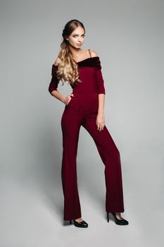 Full length of blonde model with long wavy hair in tail posing in fashionable elegant overall of bordo color with fur rim and bare shoulders, wearing high heels.