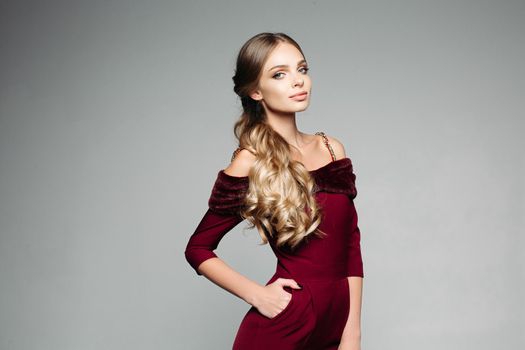 Studio portrait of attractive blonde model with long wavy hair in tail posing in trendy elegant overall with fur on bare shoulders holding hand in pocket. Isolate on grey.