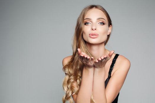 Studio portrait of an attractive blond woman with long thick hair and professional make-up wearing an elegant silver blouse, an air kiss on the camera on a gray background. Isolate. The concept of beauty.