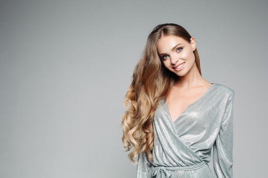 Long haired blondie woman wearing in shiny silver outfit smiling at camera. Gorgeous girl with perfect skin, curly hair, professional make up, after beauty salon posing at studio. Fashion and beauty.