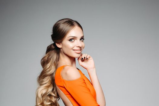 Portrait of long haired blonde woman wearing in orange outfit, smiling and touching her face by fingers. Girl with perfect skin and professional make up posing at grey studio background.