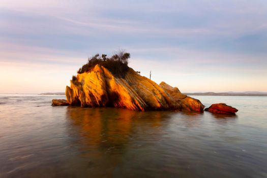 Small rocky outcrop surrounded by water, sun hitting its surfaces and accentuating its colours and textures, in the early morning