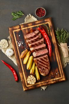 Keto ketogenic diet grilled fried beef steak, striploin, sliced potatoes on cutting board on dark brown table. Paleo food recipe with chopped piece of meat, top view, vertical