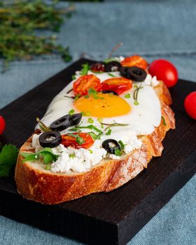 Toasted bread toast with fried eggs with yellow yolk and tomatoes, olives, sprinkled with herbs on dark wooden serving board on blue denim napkin, vertical, side view