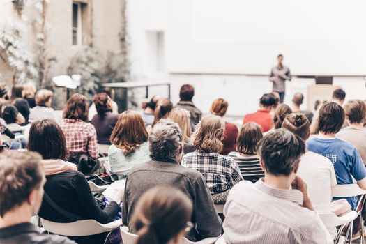 Male speaker giving presentation in lecture hall at university workshop. Audience in conference hall. Rear view of unrecognized participant in audience. Scientific conference event.