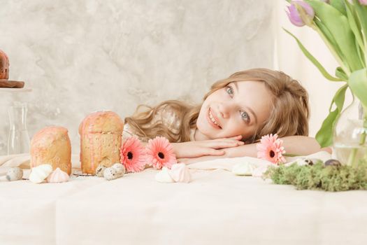 A girl with long hair in a light dress is sitting at the Easter table with cakes, spring flowers and quail eggs. Happy Easter celebration