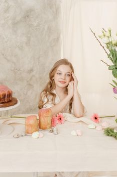A girl with long hair in a light dress is sitting at the Easter table with cakes, spring flowers and quail eggs. Happy Easter celebration
