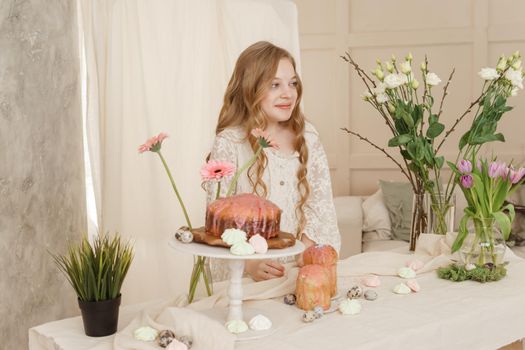 A girl with long hair in a light dress is sitting at the Easter table with cakes, spring flowers and quail eggs. Happy Easter celebration