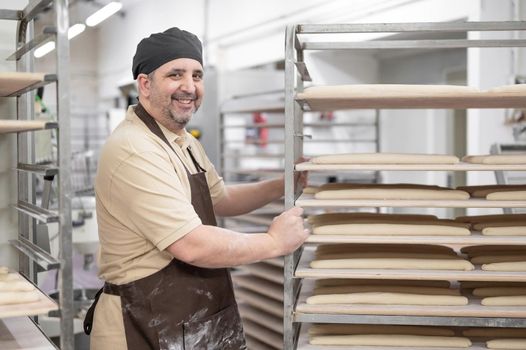 Confident baker posing with rack of fresh bread loaf at bakery. High quality photo