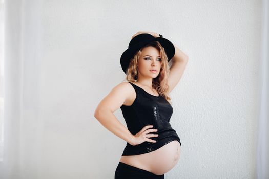 Studio portrait of attractive blonde woman with professional make up embracing her belly awaiting baby, wearing party black top and stylish black hat over white background.
