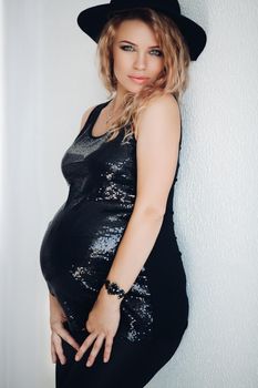 Portrait of beautiful pregnant curly blonde woman wearing in shiny black dress and hat,looking at camera over shoulder. Gorgeous female in expecting of child with makeup, hairstyle after beauty salon.