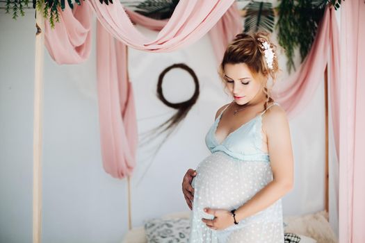 Above view for beautiful blonde pregnant woman, lying on bed, touching, holding hands on stomach, looking at camera. Pretty young mom wearing in blue expecting for her baby. Stylish home interior.