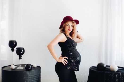 Side view of trendy lady in red hat and black top with spakling elements awaiting baby posing between two tables with modern furniture. Cutout on white background.