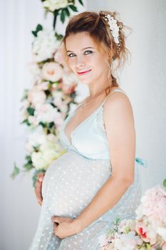 Beautiful and sensuality pregnant woman with blue eyes and hairstyle, wearing in lingerie dress. Smiling young mom expacting baby, embrasing stomach and smiling at camera. Flowers on background.