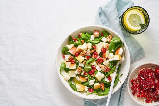 Fruits salad with nuts, balanced food, clean eating. Spinach with apples, pecans and feta, garnished with pomegranate seeds in bowl on table with white tablecloth. Copy space
