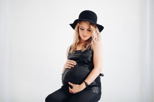 Studio portrait of attractive fashionable pregnant woman in sparkling black top and hat embracing her belly over white background.