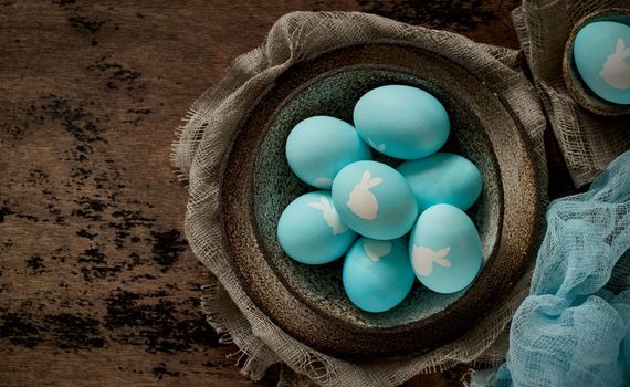 Unusual Easter on dark old background. Ceramic brown bowl with blue eggs with rabbit. Darkness, rays of sunlight shine on eggs. Concept of new life, rebirth. Rustic style. Top view, copy space