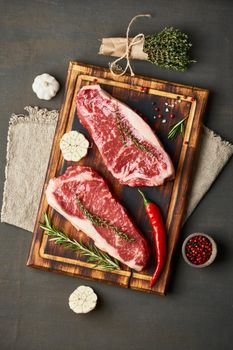 Big whole piece of raw beef meat. Seasoning steak with salt, thyme, garlic. Vertical. Two striploin on wooden cutting board on dark brown background.