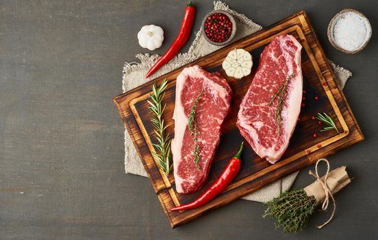 Big whole piece of raw beef meat, striploin on wooden cutting board on dark brown background. Seasoning steak with salt, thyme, garlic. Top view, copy space