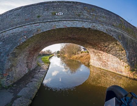 View from bow of narrowboat showing English rural countryside scenery on British waterway canal with old stone road bridge