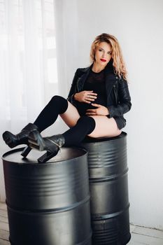 Studio portrait of pregnant lady in stylish leather jacket, knee socks and high heels posing on black containers embracing belly and looking at camera.