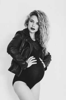 Black and white studio portrait of beautiful curly pregnant girl posing, holding stomach by hands and looking at camera. Gorgeous mom waiting for baby. Female with makeup, wearing in leather jacket.